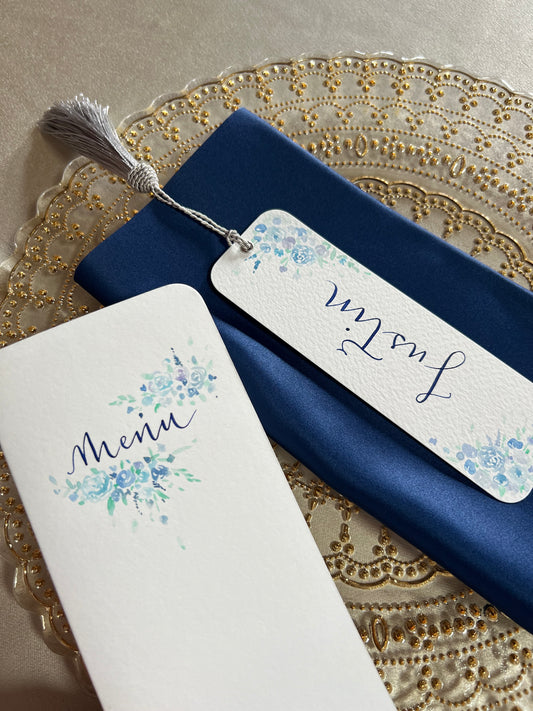 Close of pictures of wedding breakfast menu and personalised place card bookmark. Each hand painted with florals in blue hues and finished with handwritten words ‘menu’ and ‘Justin’ in navy ink calligraphy