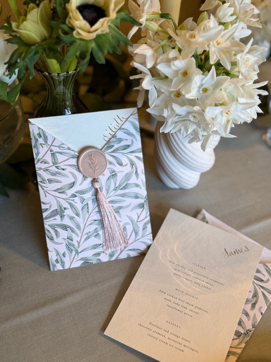 Wedding invitation with floral design and tassel, placed on a table with flowers in the background.
