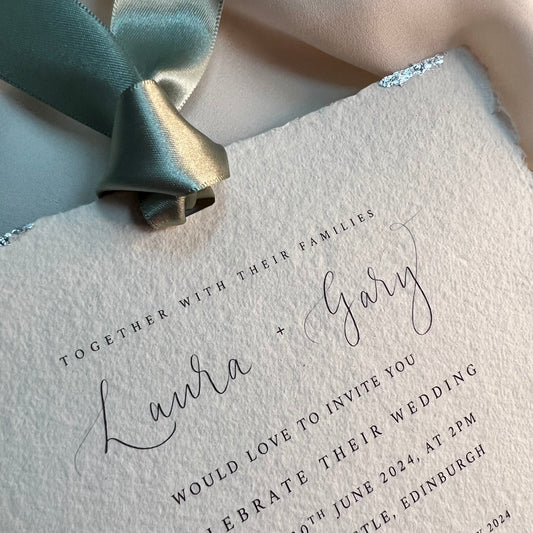Close up of wedding invitation soft edged handmade paper, green satin ribbon, silver leaf around edges