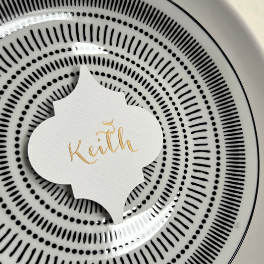 black and white charger plate with white Moroccan tile shaped place card with gold ink calligraphy reading Keith
