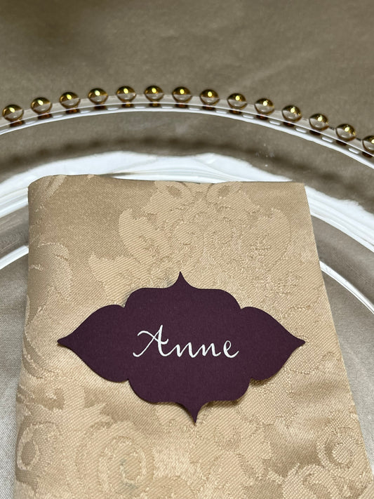 a table name card with the name handwritten in calligraphy with white ink. The card is purple and the shape is abstract but with a flowery feel. table name is lying on a napkin which is on top of a elegant silver plate.