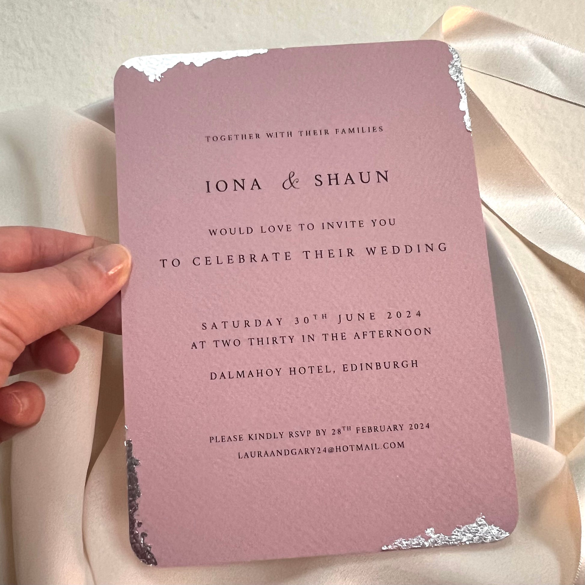 hand holding dusty pink wedding invitation with silver foil detail around edges 