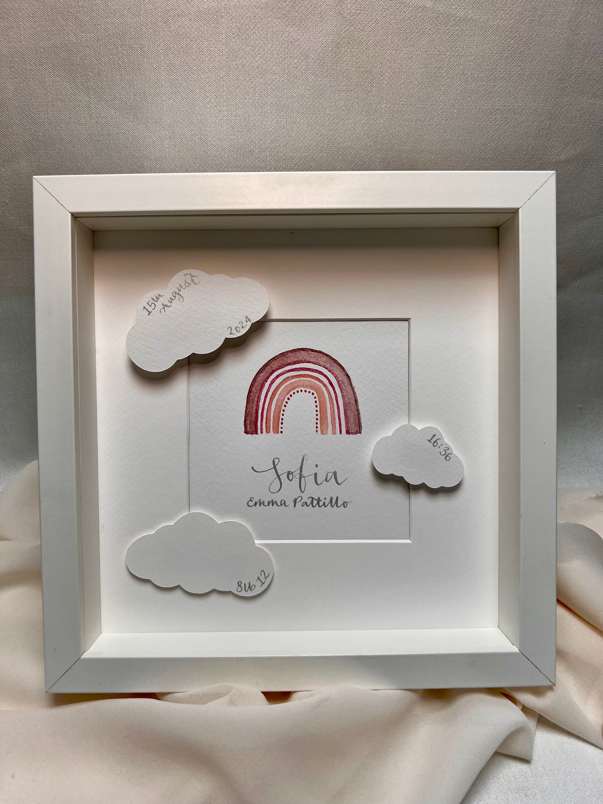 Framed watercolour rainbow painting with baby name in silver ink calligraphy and details in clouds including date of birth, time of birth and weight of baby. 