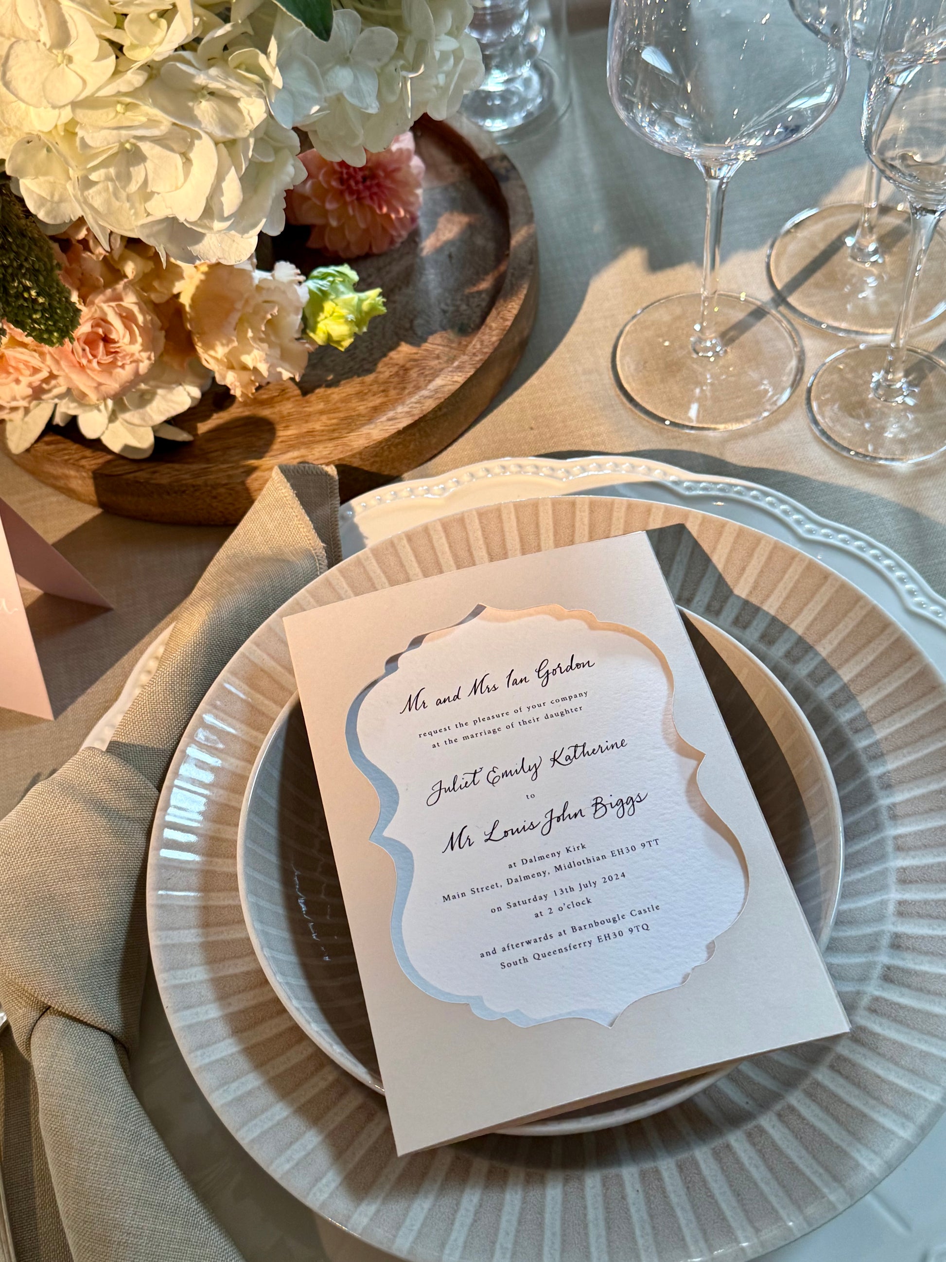 Table setting with a place card on a plate, flowers, and wine glasses.