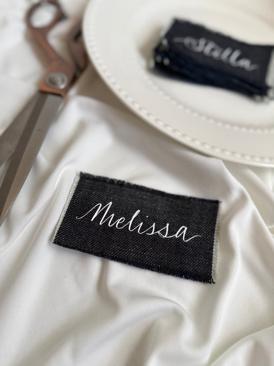 Dark denim guest names with 'Melissa' in white calligraphy writing. Fabric scissors in background 