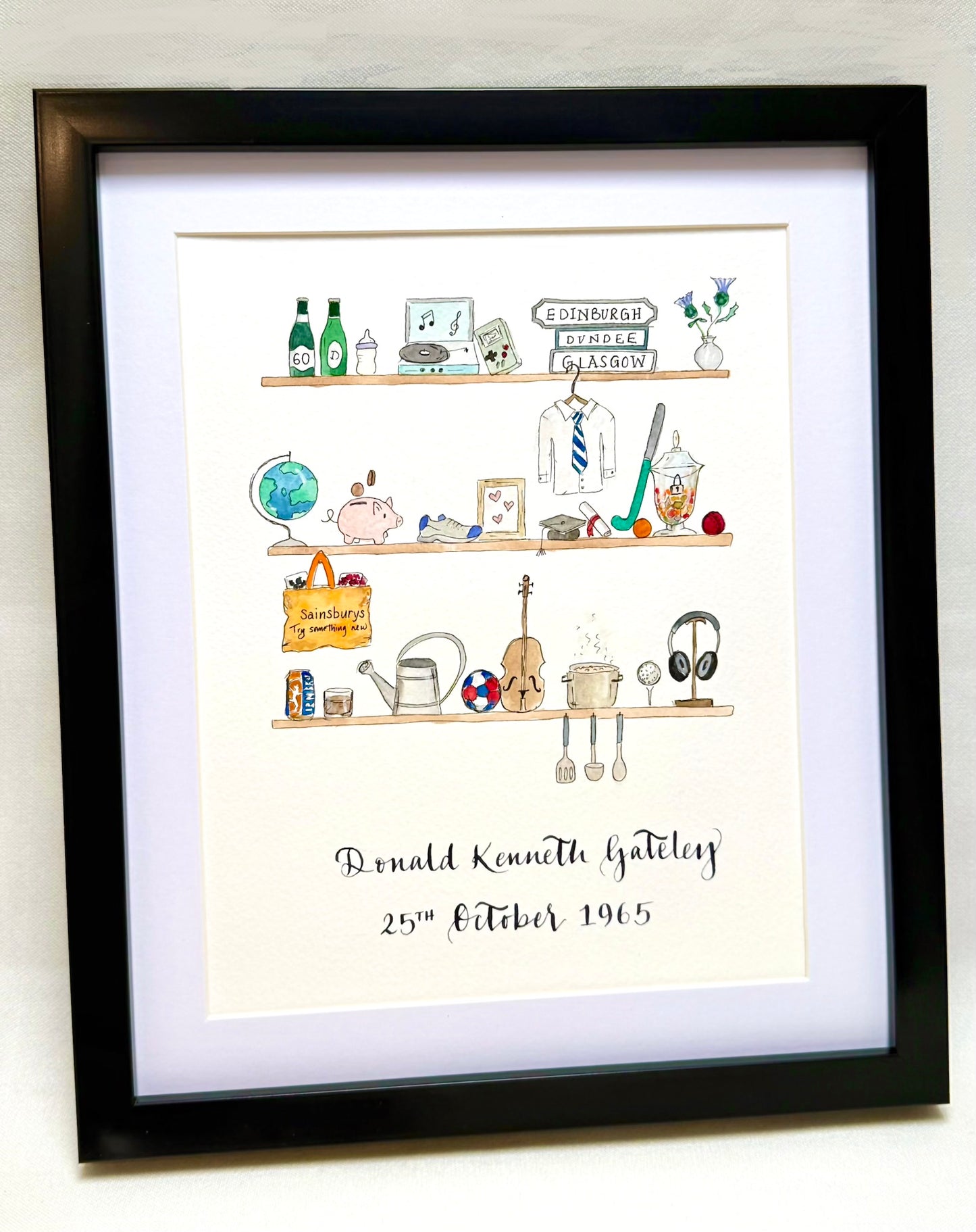 Personalised illustrated frame featuring three hand-drawn shelves with watercolour details, each showing a collection of meaningful objects such as bottles, books, musical instruments and flowers. The artwork includes handwritten calligraphy with the name ‘Donald Kenneth Gateley’ and the date ‘25th October 1965’ beneath, framed in black with a white mount.