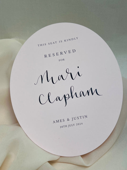 Palest pink oval card placed on cream fabric, printed with names and date of wedding, finished with handwritten guest name in calligraphy. Perfect for any wedding ceremony 