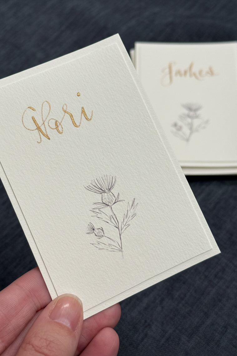 A hand holds a cream-colored textured paper card featuring gold foil script reading 'Hori' and a delicate gray botanical illustration. In the background, a stack of similar cards is visible on a dark textured surface.