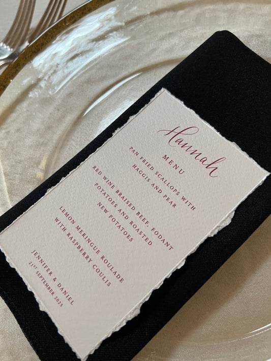 Elegant wedding place setting with a handmade deckled-edge menu card in soft ivory, printed in a berry pink ink. The menu rests on a black napkin atop a white textured plate, with gold-rimmed cutlery to the side. The menu lists pan-fried scallops, red wine braised beef, and lemon meringue roulade, with the names Jennifer and Daniel and the date 21st September 2025 printed at the bottom
