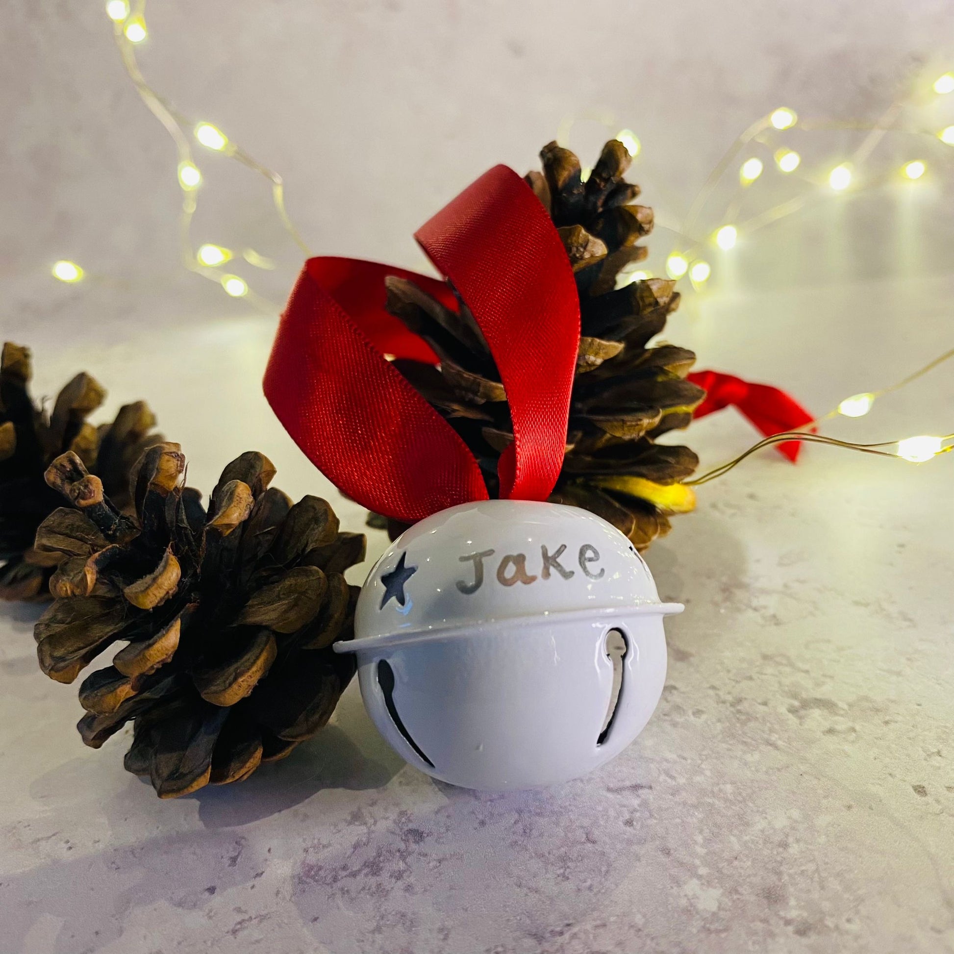 White christmas bell decoration personalised with Jake. pinecones in background and red ribbon
