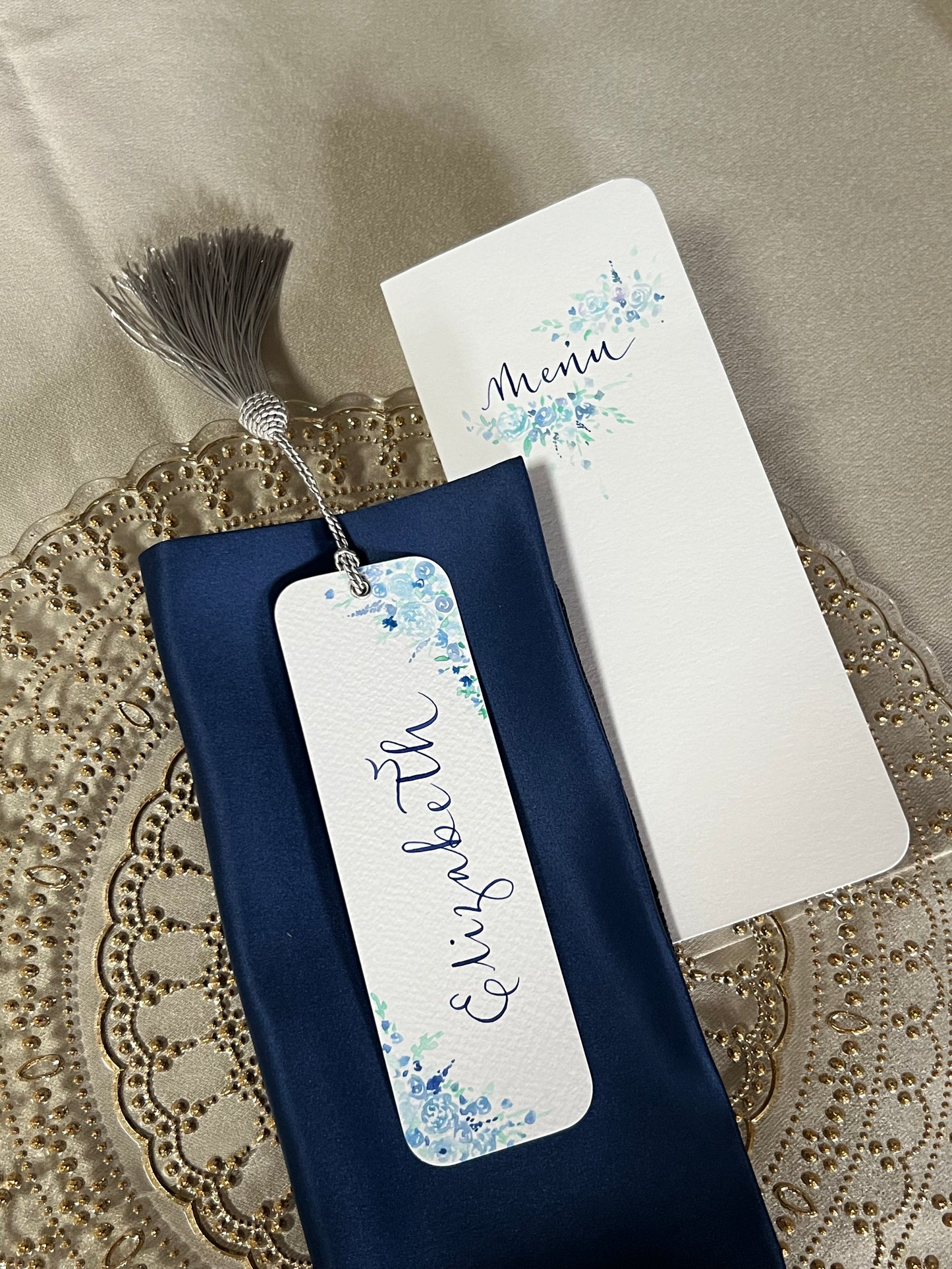 Ornate gold and glass charger plate with navy napkin, tall hand painted menu and stunning hand painted bookmark with blue watercolour florals and silver tassel 