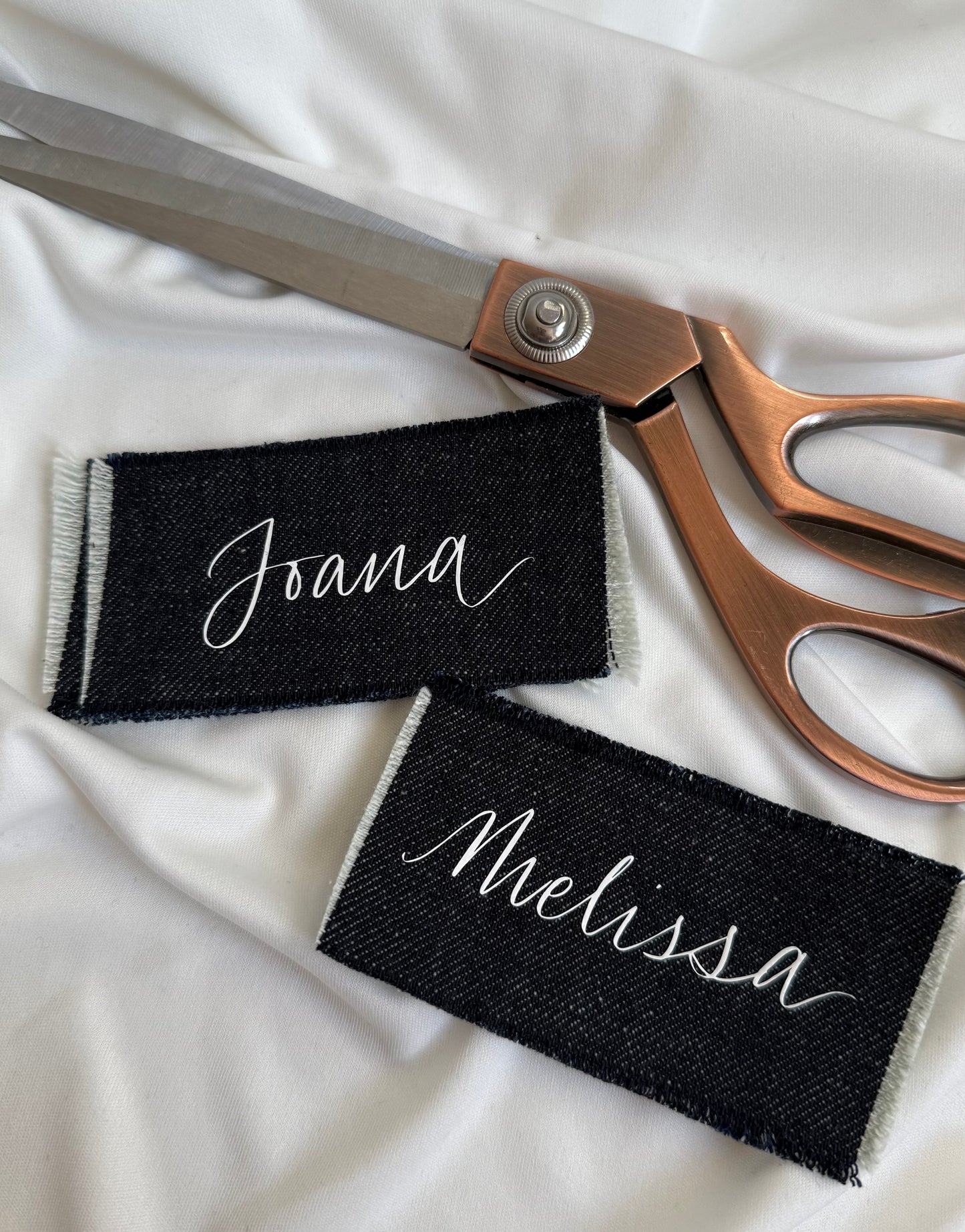 Two indigo denim name cards with 'Joana' and 'Melissa' and a pair of scissors on a white background.