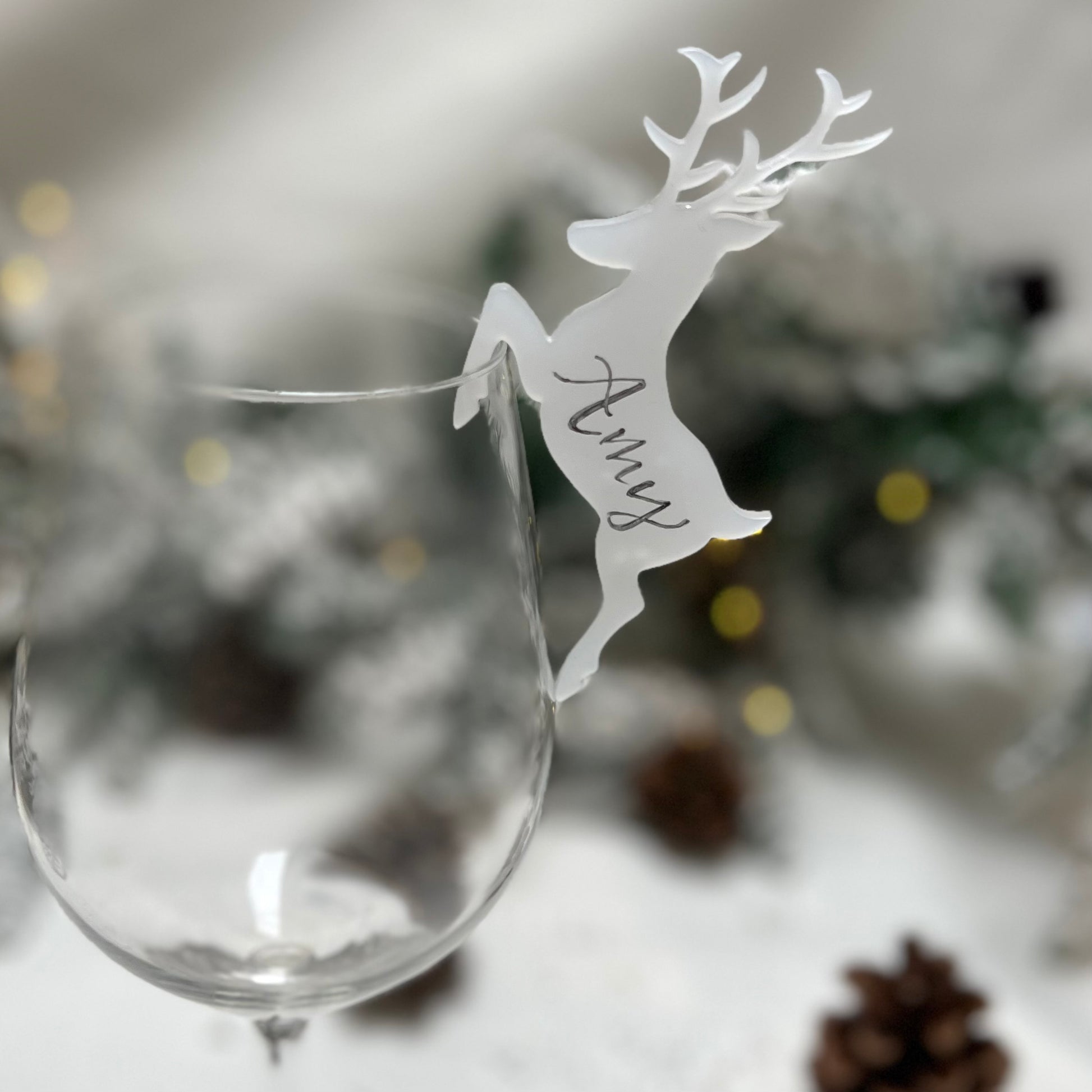 A glass showcasing a detailed deer design, made of flat, frosted acrylic. The deer is personalised with the name "Amy" handwritten on the side. the backround is blurry but appears to be snowy with acorns and Christmas trees.