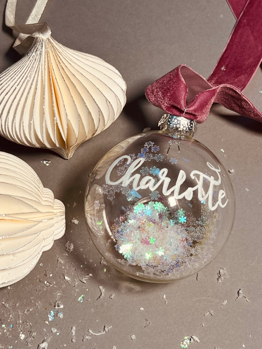 Personalised Christmas calligraphy glass bauble with dusty pink ribbon, finished with handwritten name in white ink calligraphy. Inside the bauble is a sprinkle of iridescent snowflake confetti