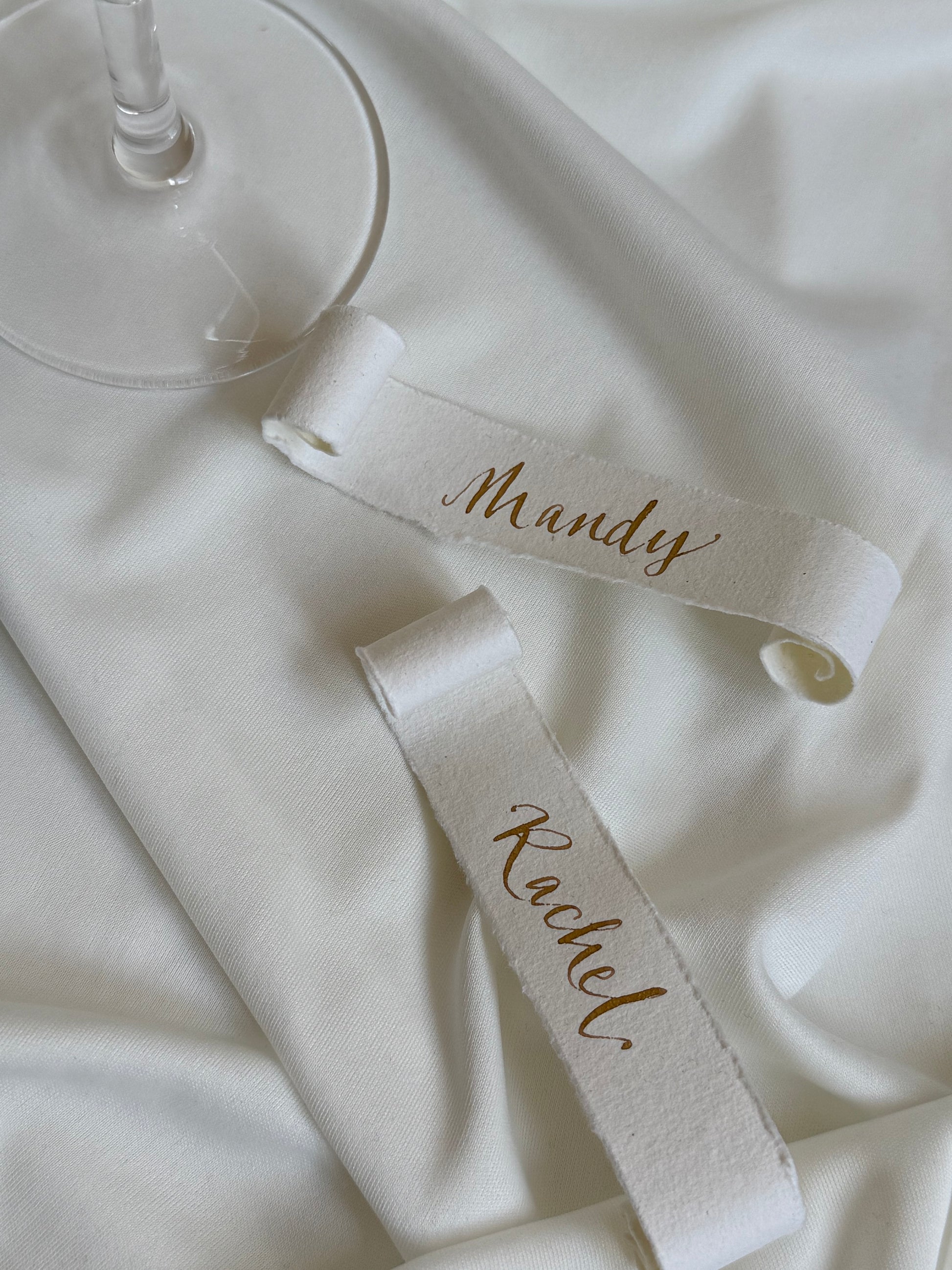 Two cotton paper place cards with names 'Mandy' and 'Rachel' in gold ink calligraphy. On a white fabric background.