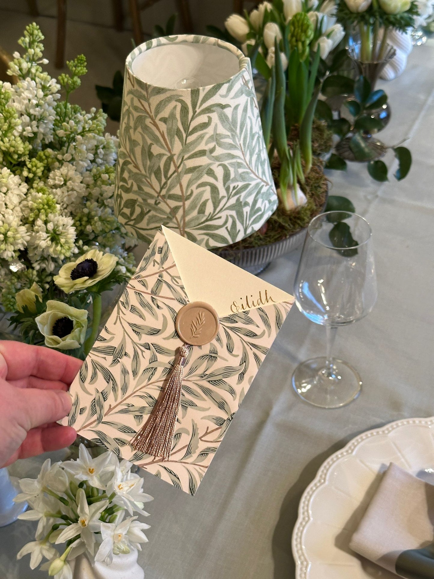 Decorative envelope with tassel and wax seal held over a table with flowers and a lamp.