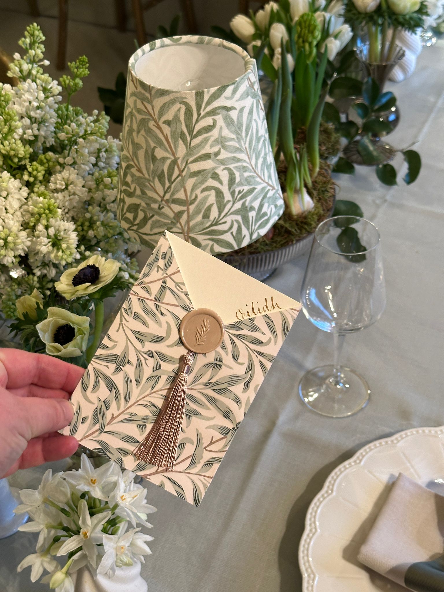 Decorative envelope with tassel and wax seal held over a table with flowers and a lamp.