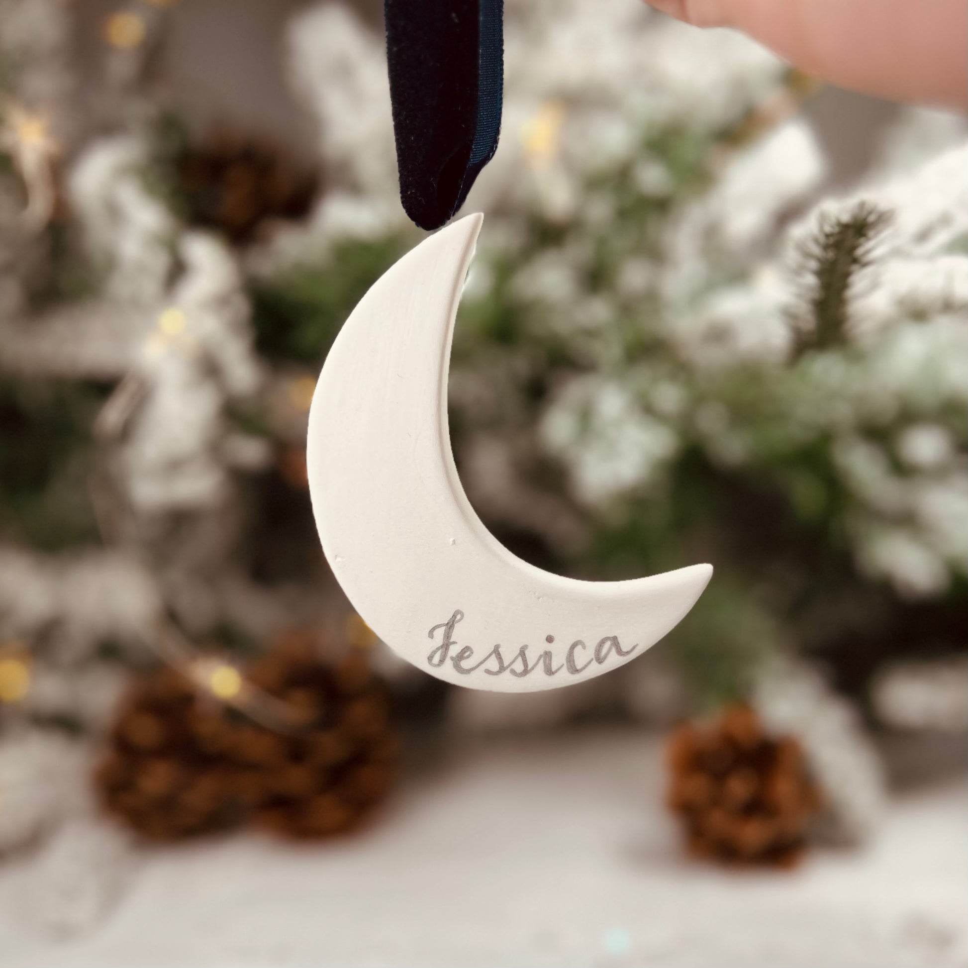 background of unfocused winter greenery, hanging ceramic moon with handwritten name, hanging with navy velvet ribbon