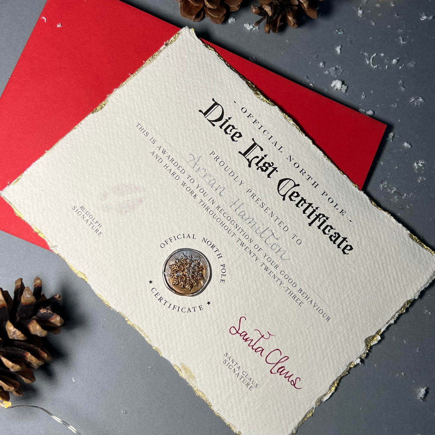 A bespoke Nice List certificate, with golden, frayed edges and a snowflake wax seal from the North Pole awarded to a child, signed by Santa Claus and Rudolph, placed on a red envelope with pinecones and festive lights in the background.