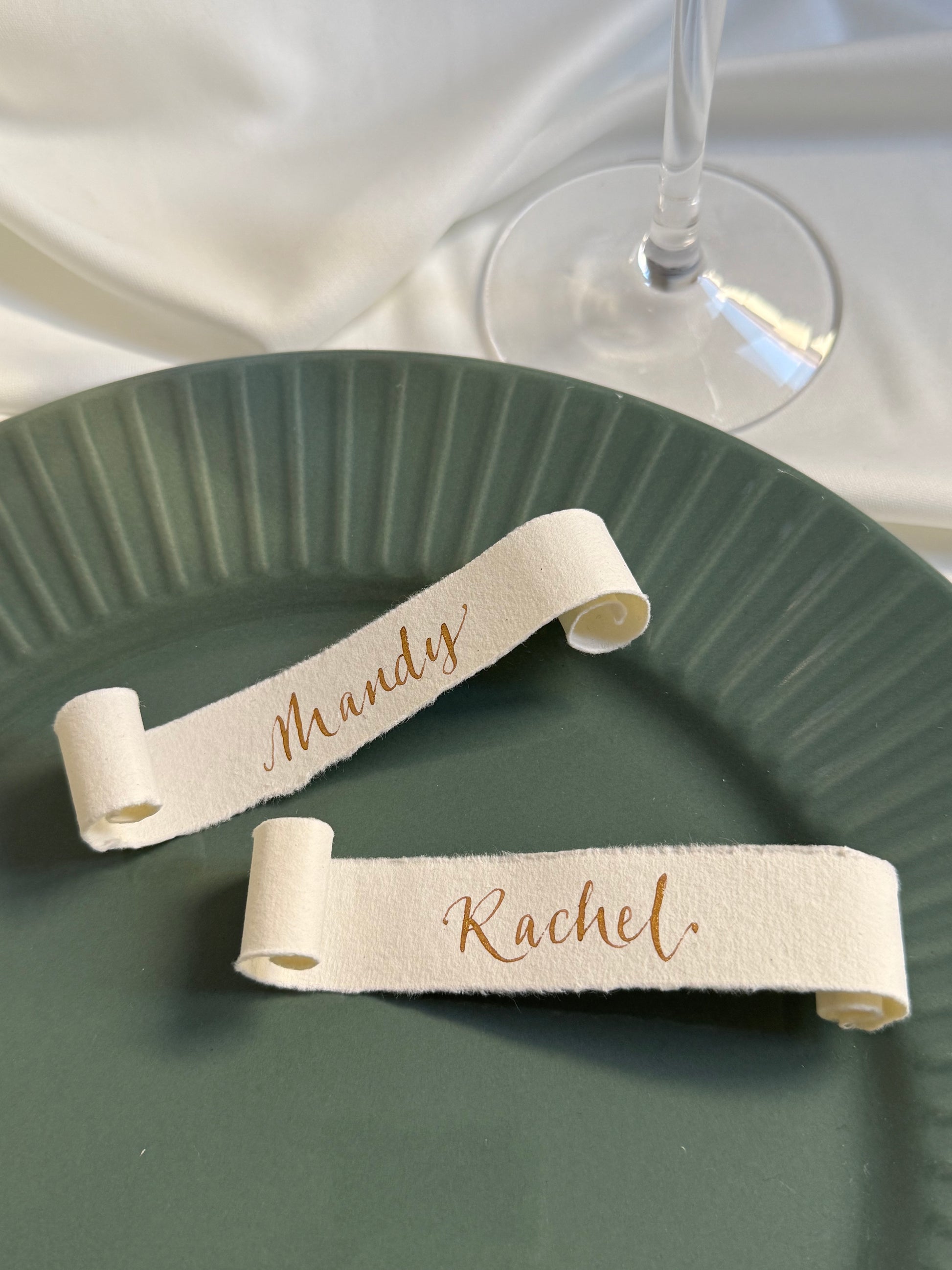 Two cotton paper scroll name tags on a green plate with a glass of wine in the background