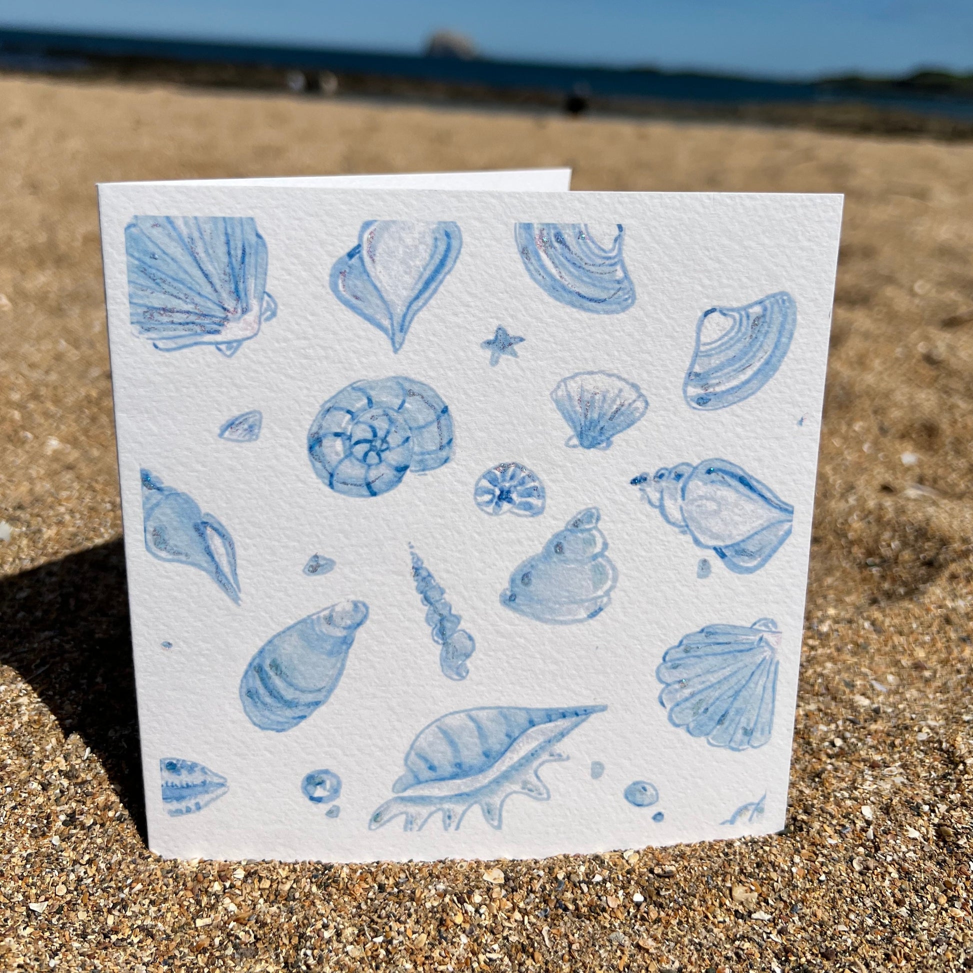 Printed art card - various watercolour shells all in blue tones sitting on sand with beach in background