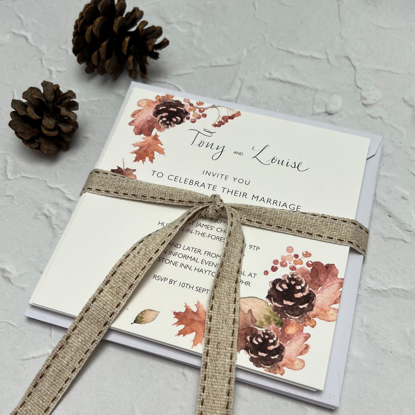 A bespoke wedding invitation with an autumn theme, adorned with pine cones and leaves, perfect for a woodland-themed event. the background is white with large texture and in the top right are two real pinecones.
