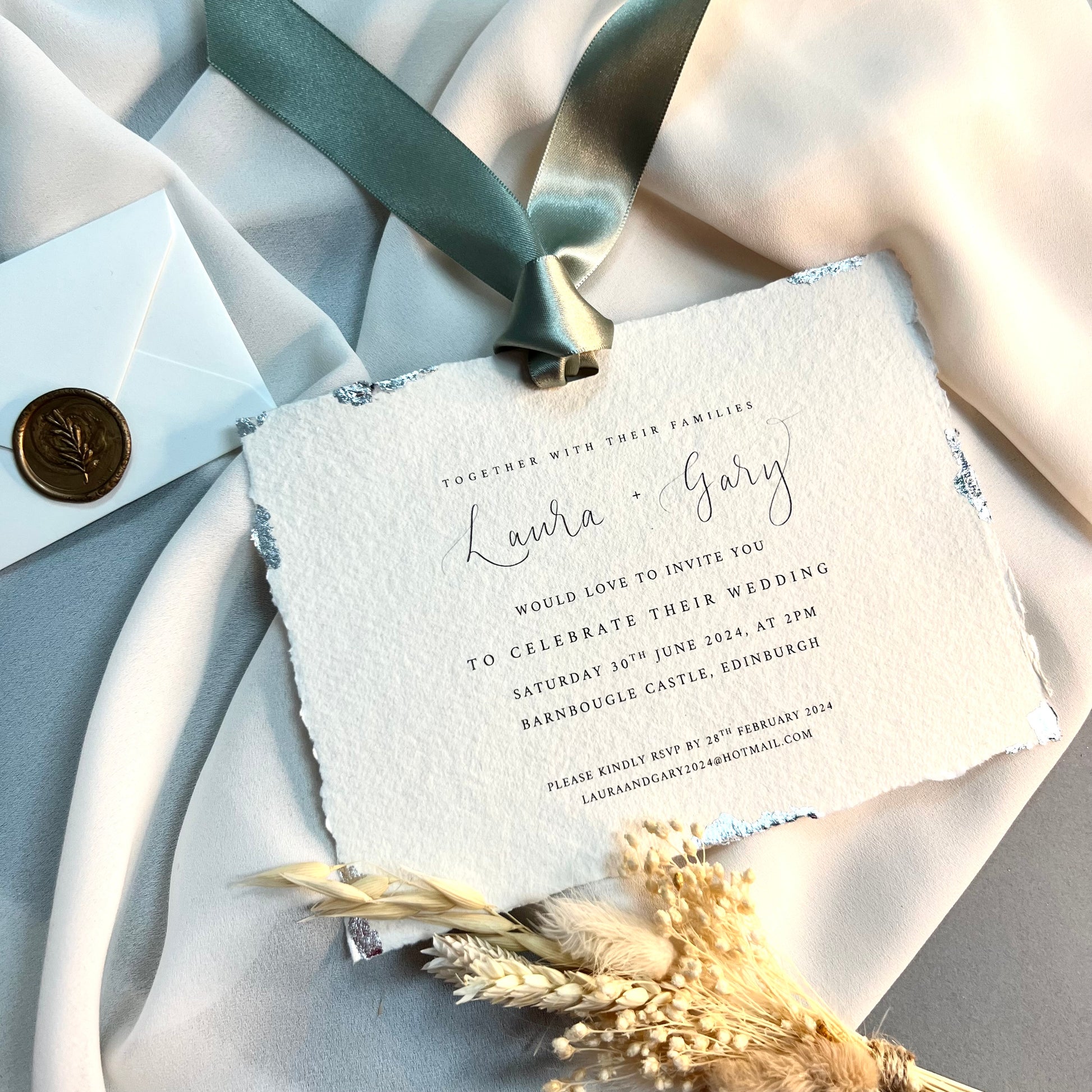 Rough edged paper with silver detail around edges, invitation printed on paper and sage green satin ribbon at top. Invitation on ivory fabric with dried grass and wax sealed envelope