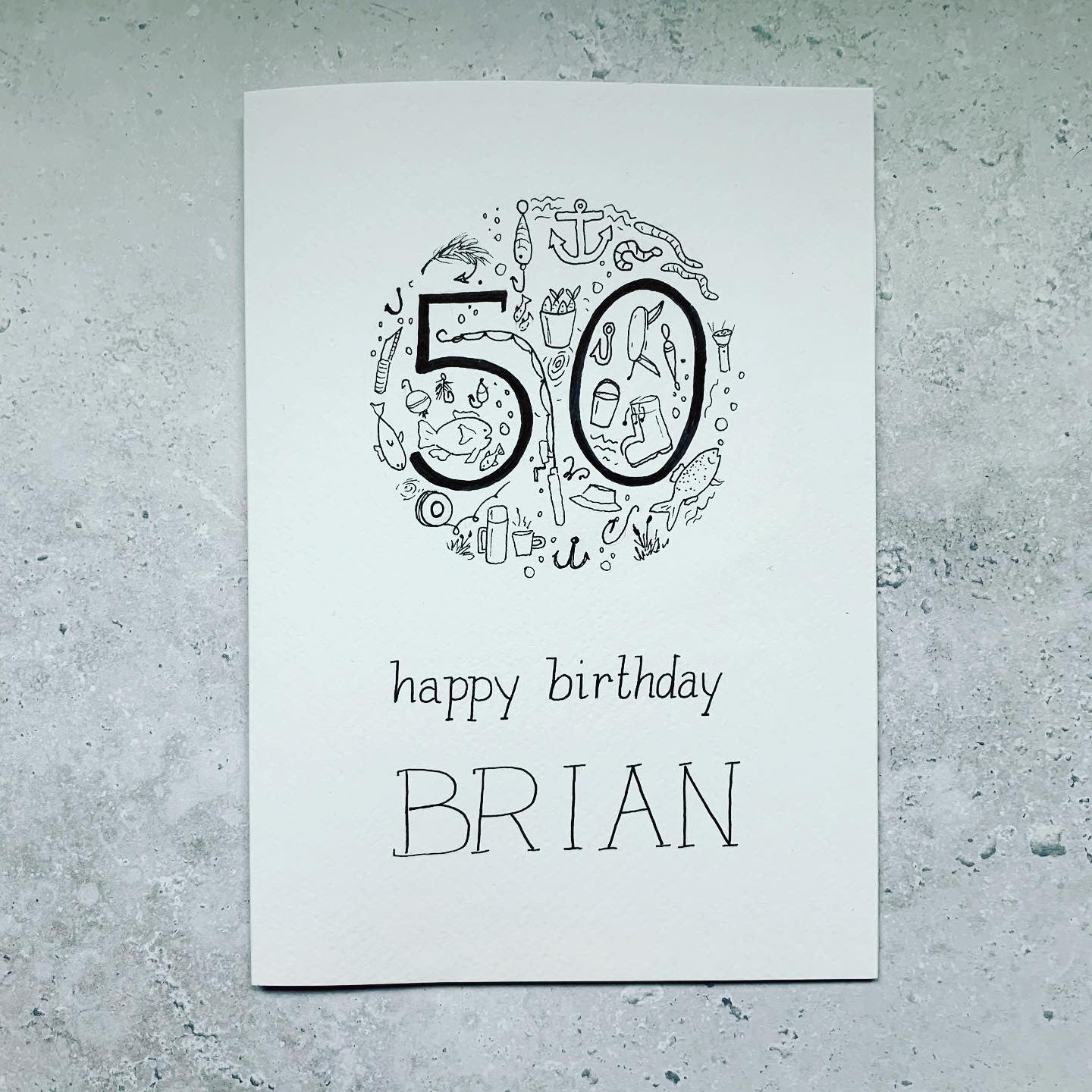 Image of full card on a background. Bespoke, handmade 50th birthday card personalised with fishing iconography such as fishing rod, fishing reel, anchor, waders, flask, bait and fish making a circle shaped design. Black and white artwork. “Happy birthday Brian” handwritten.