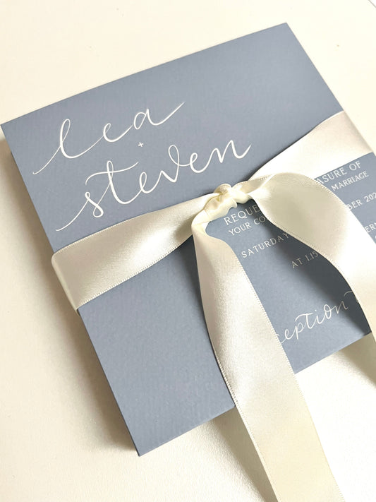 A bespoke blue wedding invitation with white handwritten calligraphy, elegantly adorned with a ribbon, showcasing a beautiful design for a special occasion.