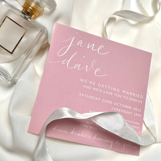 bespoke, pink square invite with bride and groom names handwritten in calligraphy. invite is elegantly lying on fabric with perfume and ribbon around .