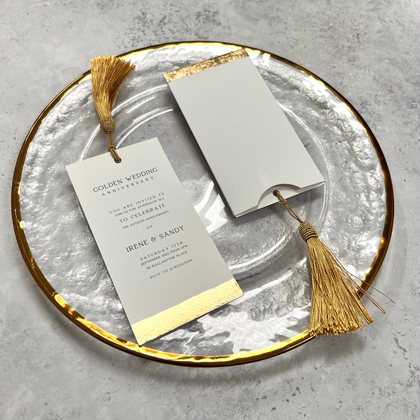 gold foiled wedding invitation - rectangular card in portrait with a golden tassel tied to the top and writing down the centre of the card in black ink, there is a strip of gold foil down the bottom edge of the card. the invitation is laid out on on a glass plate with another invite, still in its envelope sitting beside.