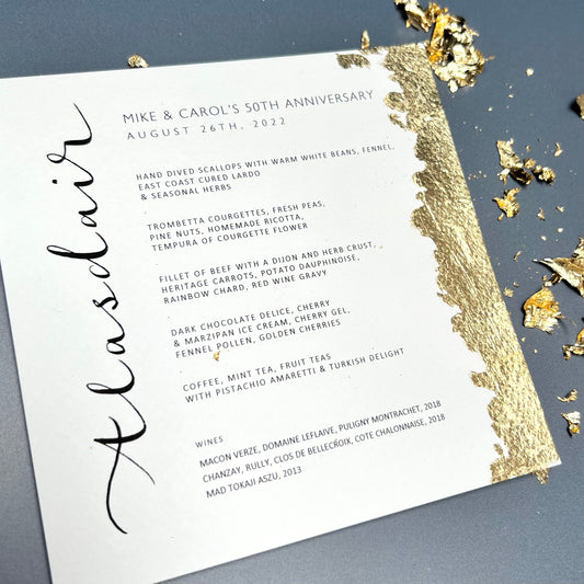 A menu card with guest name hand written in calligraphy , gold leafed on the right side of the menus with loose gold leaf coming off the side.