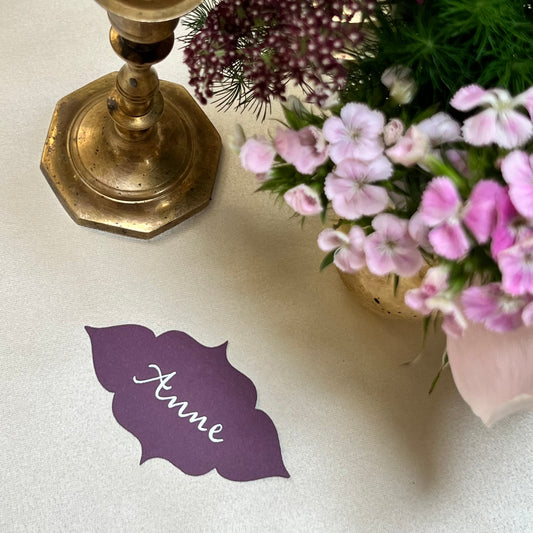 a purple table name with the name handwritten in calligraphy using white ink. the shape is abstract but with a flowery feel. the background is white fabric and has a golden candle and flowers lying beside the table name.