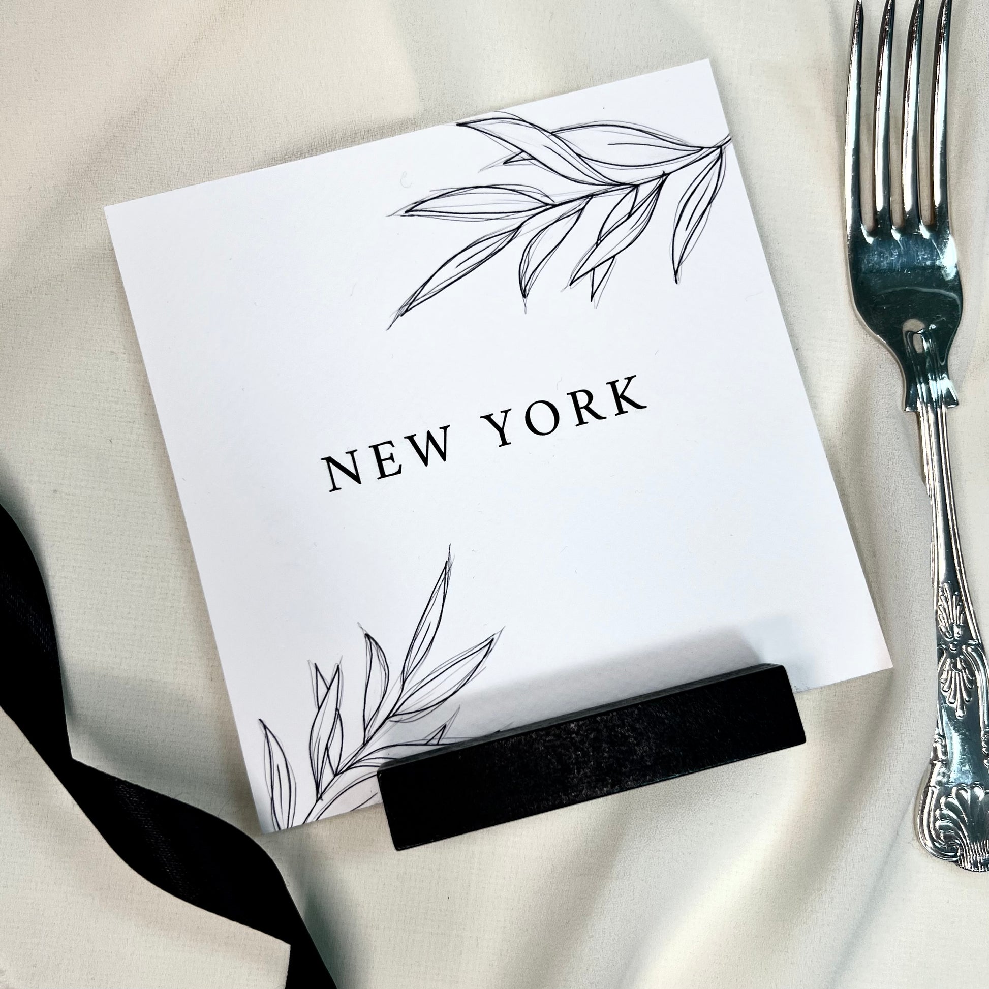 table name, square black and white card with black holder, sketch of foliages. black ribbon to left hand side and traditional silver folk to right side