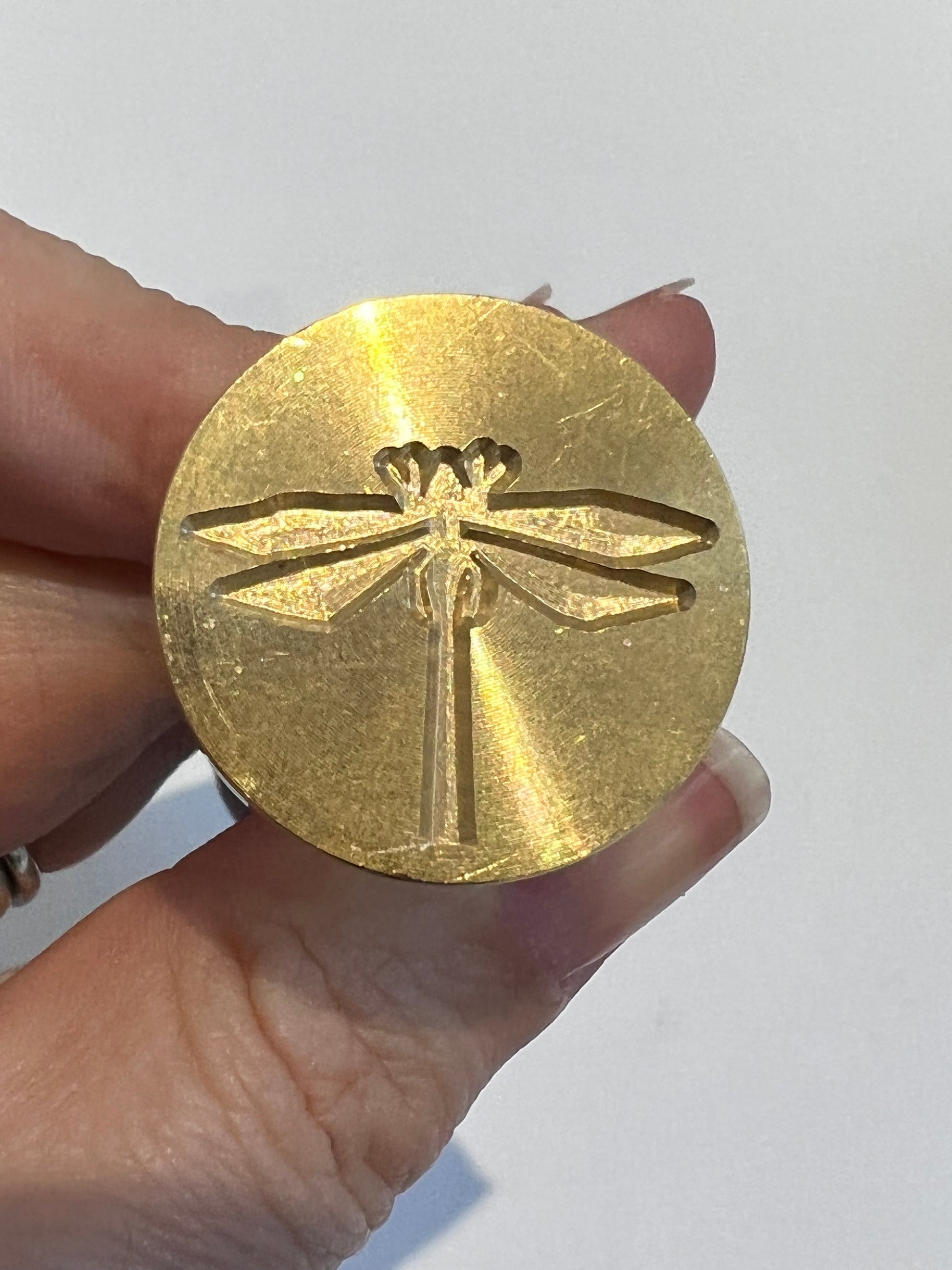 Birdseye view of a wax seal face up with dragonfly design imprinted, hand holding the handle of the wax seal and a white, uniform background.