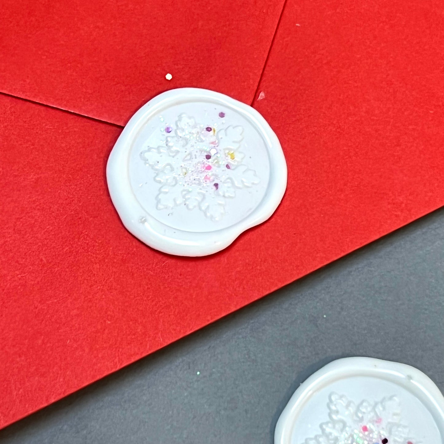 close up image of glitter wax seal on red envelope. the wax seal has a white snowflake design. to the of the image there is a wax seal without an envelope.