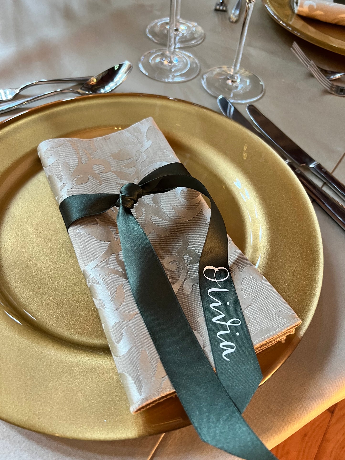 Wedding table set up with gold charger plates, damask napkins flat folded with moss green ribbon tied around and guest name on ribbon in white reading Olivia 