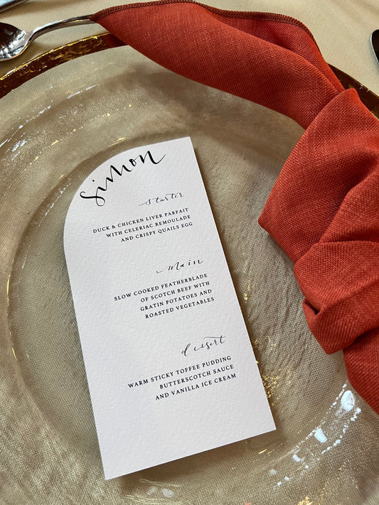 Modern curved menu on white card with black ink calligraphy name. Sitting on a glass charger plate with gold edge and burnt orange croissant knot napkin 