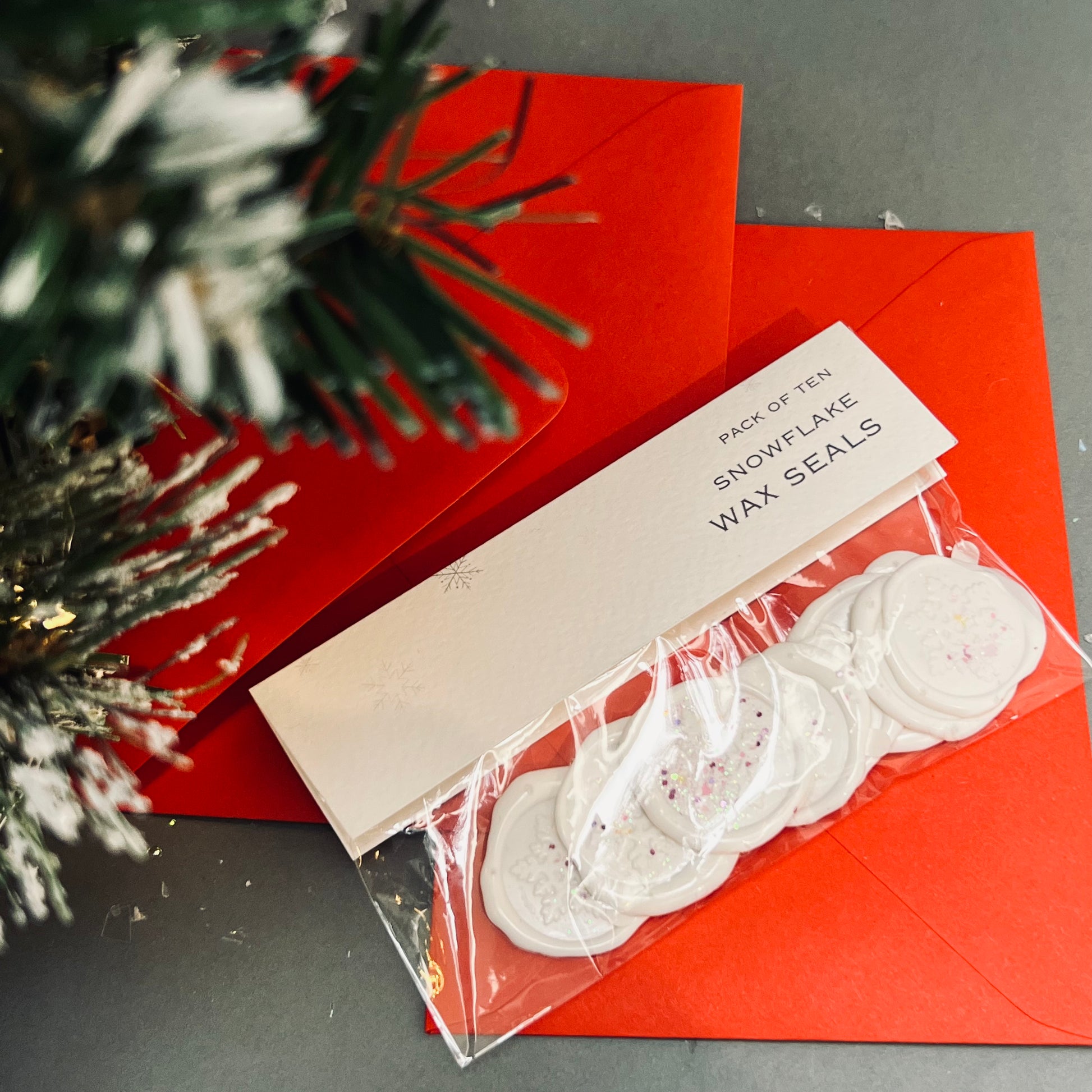 an image of handmade, Christmas themed, white snowflake wax seals in a packet which has text telling us they are wax seals, behind the packet are two red envelopes and we can just see a Christmas tree to the left side of the image.