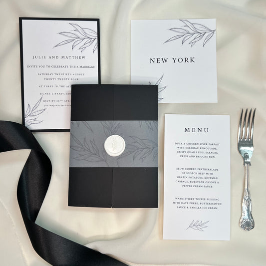 Styled photo, background of cream fabric with stationery on top, black and white invites, table name, menu with a fork to one side and black ribbon to other