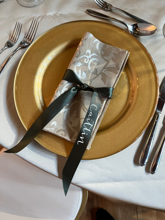 moss green ribbon tied around a damask napkin on a gold charger plate, ribbon finished with guest name in white reading Caitlin