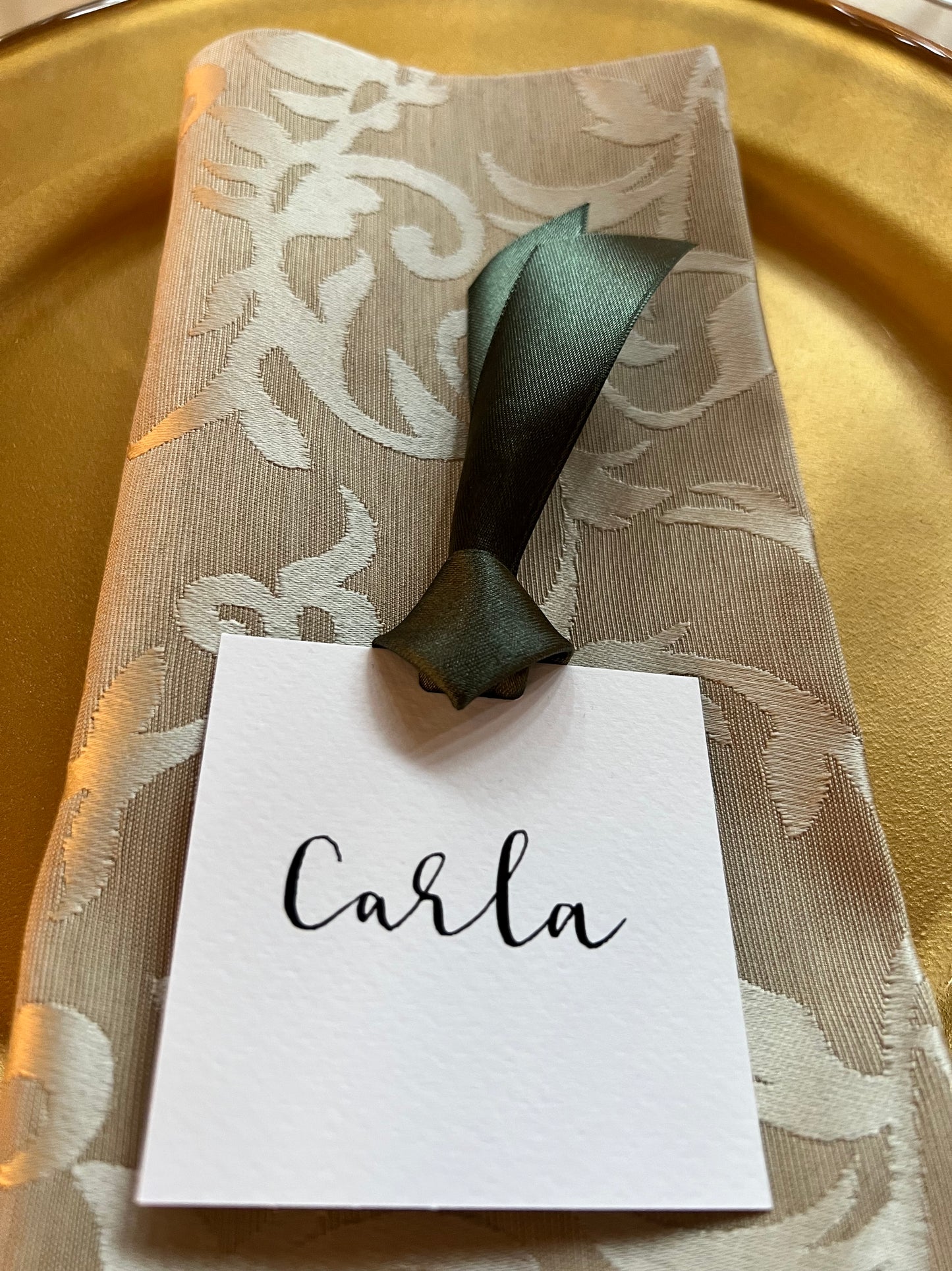 Square white card with guest name handwritten in black in calligraphy, ribbon tied to top of card. Card sitting on damask napkin, on gold charger plate.