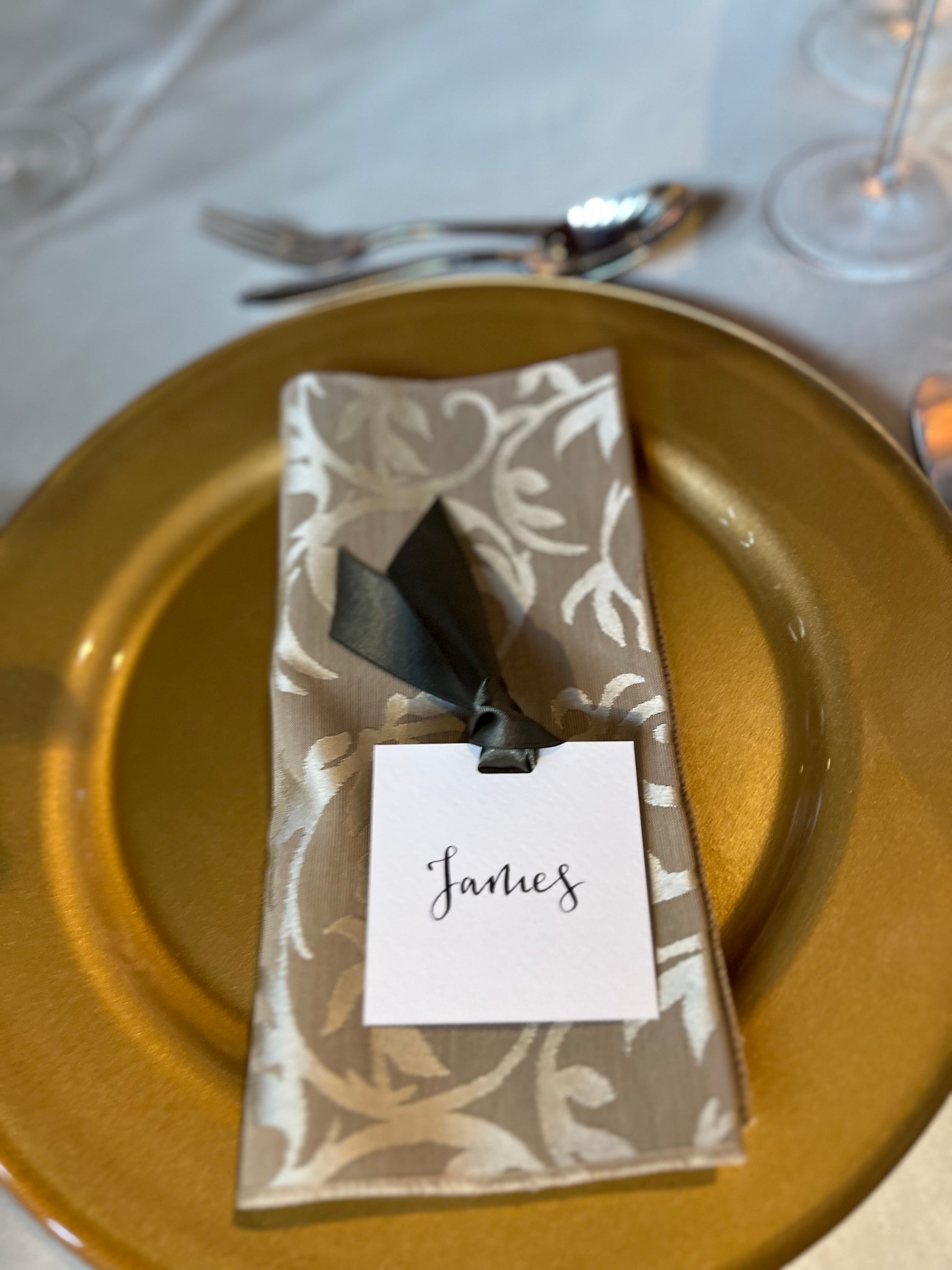 guest name place card on napkin. Gold plate with white square card and moss green ribbon