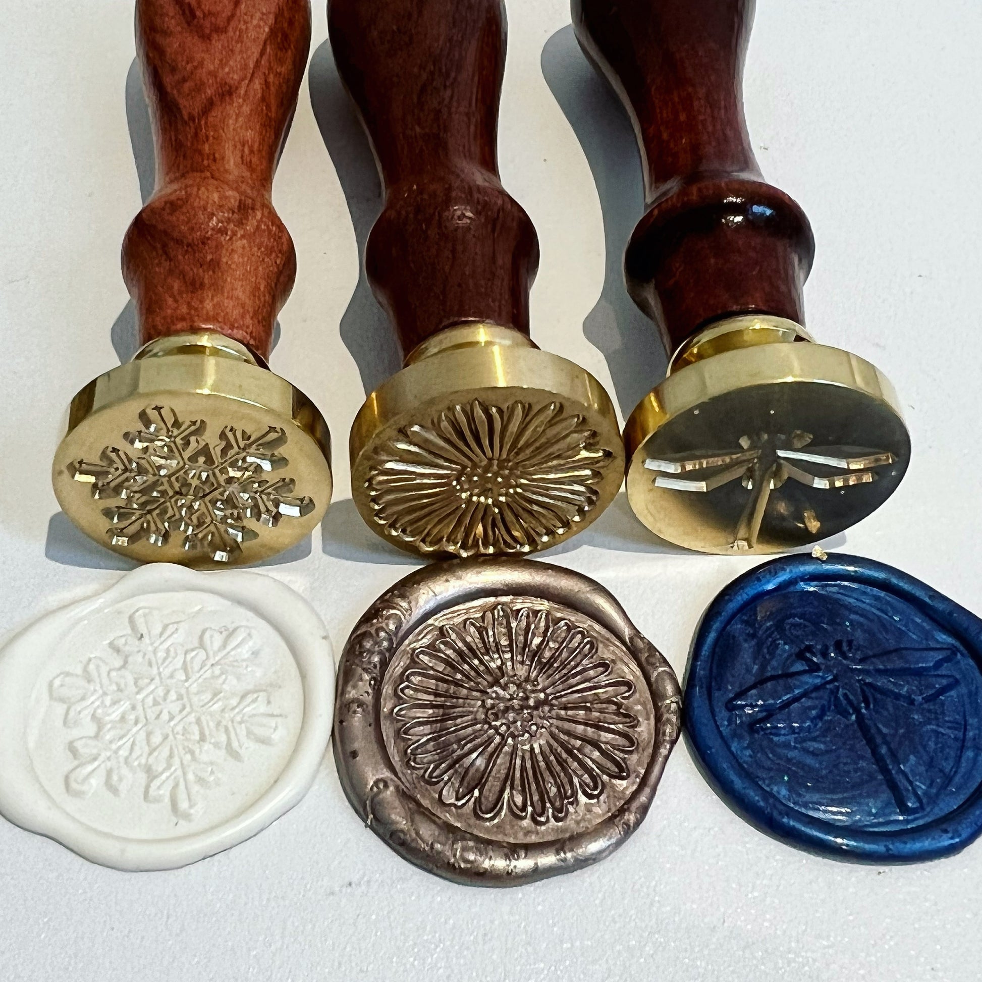 3 wax seals with snowflake, daisy and dragonfly designs imprinted into them by corresponding wax stamps laid out above them