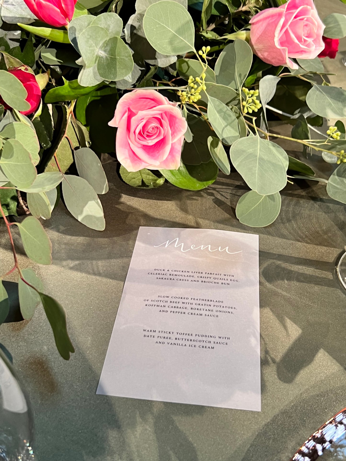 Vellum paper with printed menu and handwritten word 'menu' at top in white ink. on green table cloth with eucalyptus and roses 