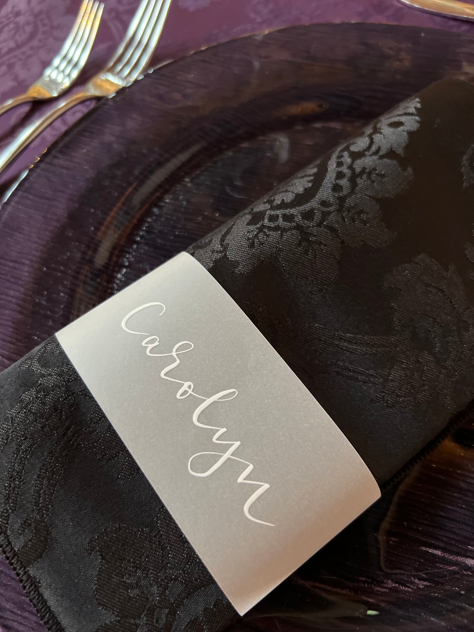 white ink calligraphy reading Carolyn, on vellum paper band which is then wrapped around a black damask napkin