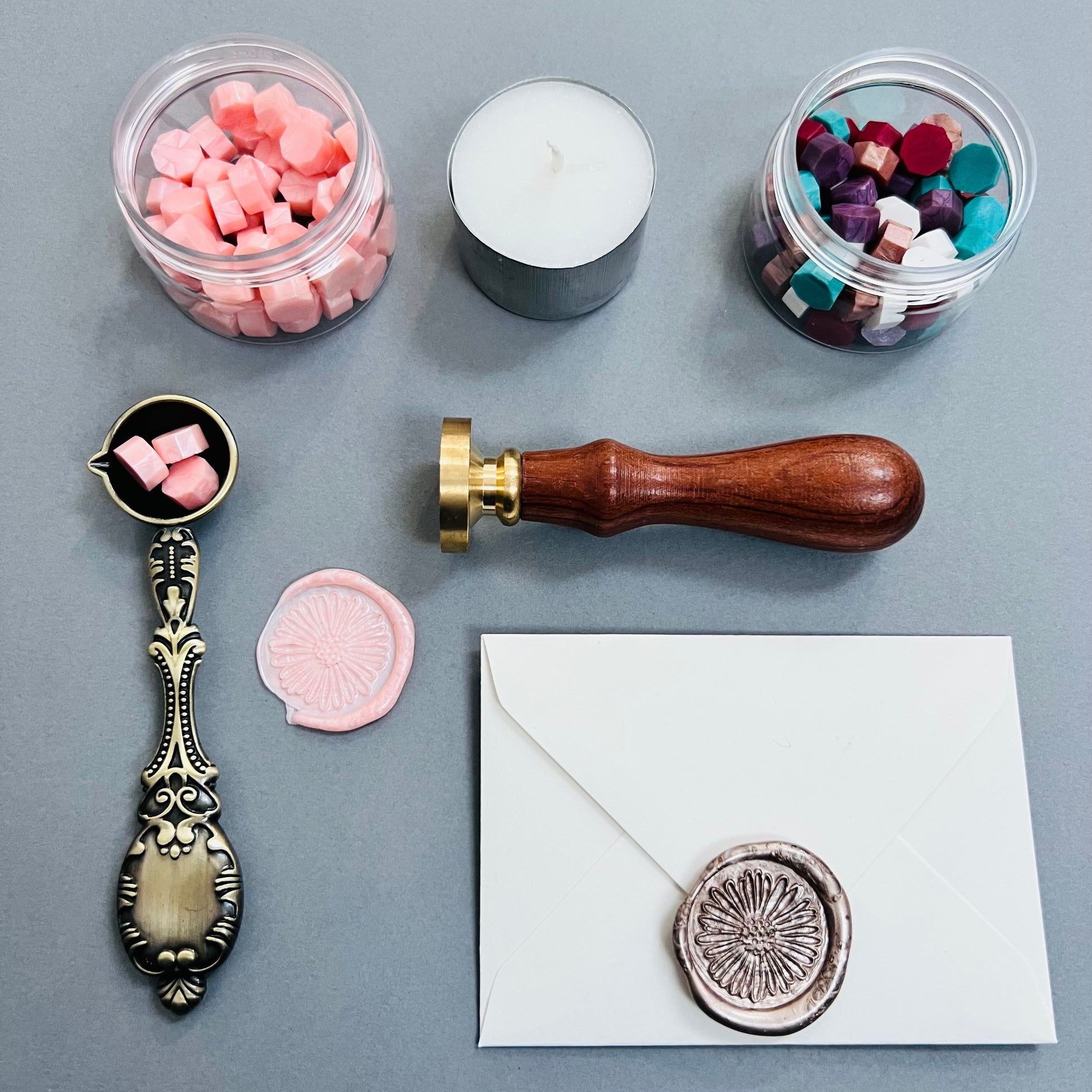 grey background with all items laid out, 2 tubs of different coloured wax pellets , wax sealing spoon, wax seal stamp, candle and an envelope with wax seal.