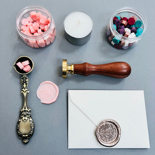 grey background with all items laid out, 2 tubs of different coloured wax pellets , wax sealing spoon, wax seal stamp, candle and an envelope with wax seal.