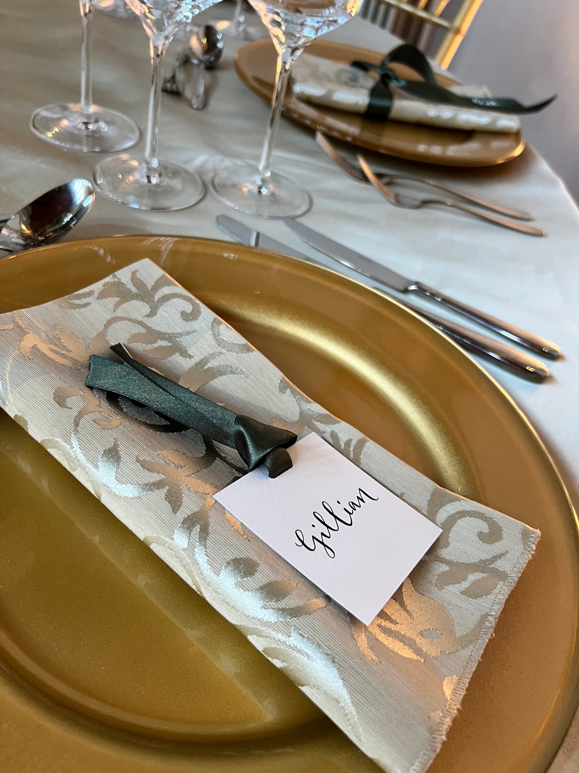 Wedding Breakfast table set up with crockery, glasses, charger plates and napkins. guest name card on top of napkin - square white card with black ink name and moss green ribbon